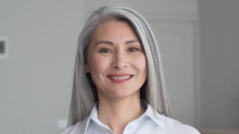 Happy smiling stylish confident 50 years old Asian female professional standing looking at camera at gray background. Portrait of sophisticated grey hair woman advertising products and services.