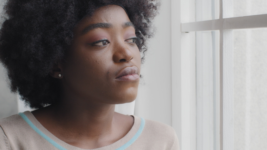 Thoughtful lonely millennial African woman look through window alone at home feeling anxious hurt offended ashamed having problem drama. Sensitive mixed race lady introvert reflect think of solitude | Shutterstock HD Video #1073348792