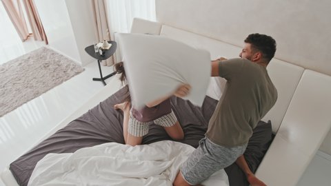 Top-view shot of young playful Latino couple having pillow fight in bed enjoying morning together at home in their modern minimalist-style apartment