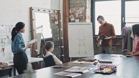 PAN shot of male fashion designer in glasses holding fabric samples and showing idea for garment drawn on whiteboard while conducting meeting with seamstresses in studio - Powered by Shutterstock - Get 15% off with code: PIKWIZARD15