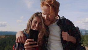 Smiling guy and girl looking video on smartphone screen during hike in mountains. Happy couple of friends using mobile phone at vacation. Handsome man hugging woman with cellphone outdoor