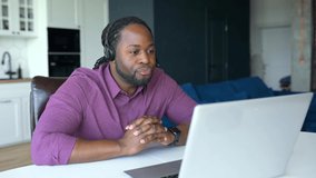 Happy African-American man with locks hair wearing wireless headset sitting at home office and greeting participants of video meeting, waving into laptop webcam, online tutor conducting video classes