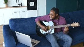 New skills concept. Concentrated African-American man learning how to play guitar with online video tutorial, a black guy watching video classes and repeats exercises sitting on the couch at home