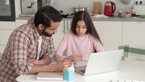 Young indian dad helping teenage daughter schoolgirl study online at home sitting at kitchen table at home using pc laptop computer. Father and kid e learning together having remote distant lesson.