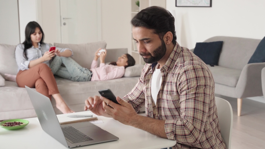 Hispanic indian dad father businessman remote working online sitting at kitchen table using laptop and phone with his wife and teen daughter at home office. Virtual home office, distance job in India.