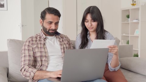 Young Indian latin family couple sitting together on sofa at home using laptop computer reading bank papers bills paying calculating loan debt taxes payments online. Wife and husband planning budget.