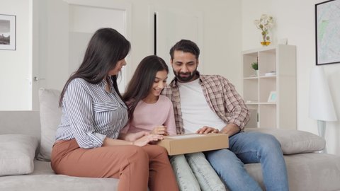 Happy excited indian family parents with teenage daughter unpacking parcel postal shipping box receiving shipment from online shop sitting on sofa in living room. E commerce online shopping concept.