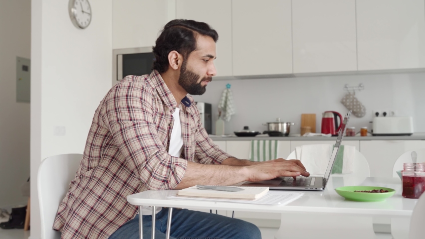 Young Indian Hispanic businessman using computer remote distance working sitting at kitchen table writing notes. Latin student studying online, having virtual digital training on laptop at home office Royalty-Free Stock Footage #1073362697