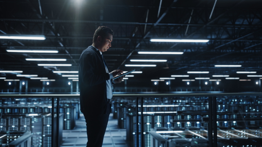Portrait of IT Specialist Using Tablet Computer in Data Center. Big Server Farm Cloud Computing Facility with Male System Administrator Working. Cyber Security, Network Protection. Full Wide Arc Royalty-Free Stock Footage #1073372453