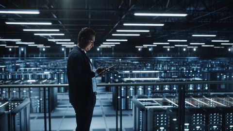 Portrait of IT Specialist Using Tablet Computer in Data Center. Big Server Farm Cloud Computing Facility with Male System Administrator Working. Cyber Security, Network Protection. Full Wide Arc