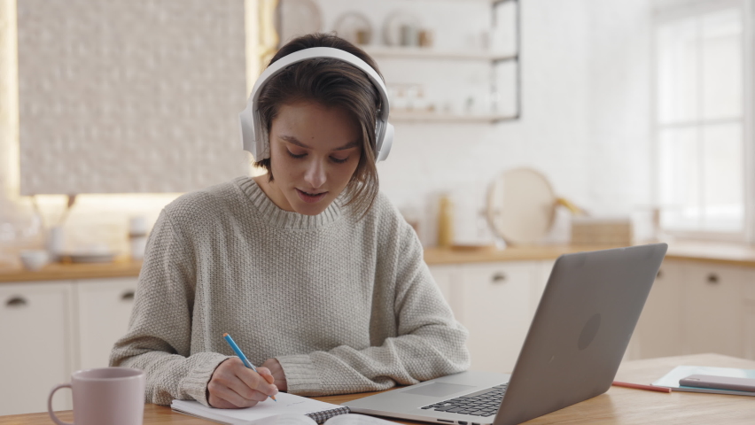 Happy caucasian woman sitting at kitchen table with modern laptop and enjoying domestic education. Pretty student in wireless headphones spending free time for studying. Royalty-Free Stock Footage #1073380178