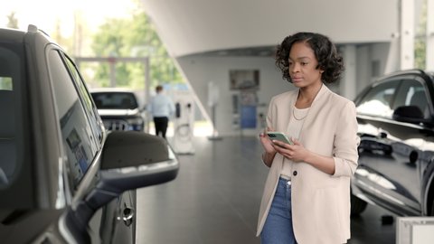 African woman with smartphone in hands standing near luxury car and examining exterior. Female client making choice at dealership before purchase.