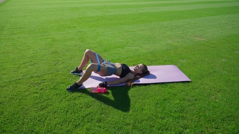 Young woman with perfect muscular body training glutes using fitness band on mat on green stadium field. Girl practicing floor bridge butt raise on grass outdoors. Workout concept.