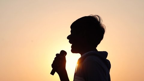 Silhouette of a kid screaming in the mic in front of the sun during the sunset. Kid singing rock song and shouting in the microphone