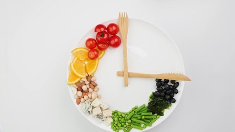 Colorful food and cutlery arranged in the form of a clock on a plate. Hand takes an olive. Intermittent fasting, diet, weight loss, lunch time concept.