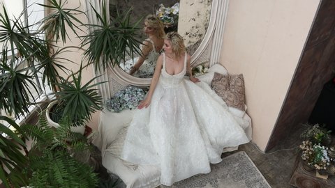 Attractive young bride blonde in beautiful wedding dress looks at herself in large wall mirror in half turn. 4K Resolution