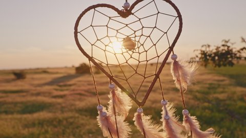 Heart shaped dream catcher with orange feathers rotate in mountains, swaying with light wind slow motion agains blue sky at sunset in summer. Amulet boho style, Indian style. Bright disk of sun, rays