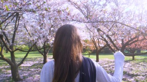Slow motion girl touching with hand playfully blossoms sakura tree. Young woman with long hair enjoys spring garden in bloom. Girl walking in Japanese Garden with blooming trees. Dreamy Soft Focus.