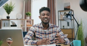 Good-looking smiling friendly 25-aged black-skinned guy sitting in front of camera during recording vlog for internet audience on beautiful home interier background