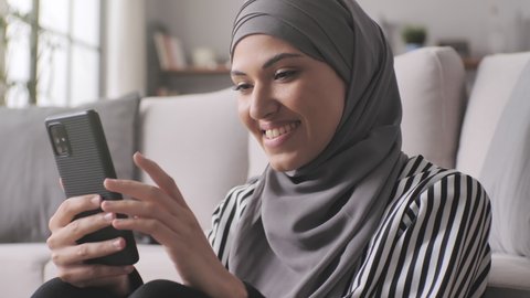 young arab muslim woman in headscarf hijab using smart phone at home,close up portrait of smiling middle eastern girl holding mobile smartphone indoors in her modern apartment