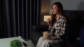 funny young woman watching TV and eating popcorn on sofa at night
