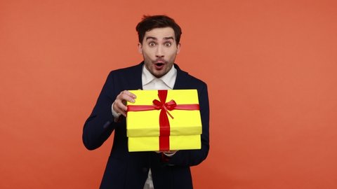 Handsome brunette man holding yellow present box in hands, shaking and interesting what inside, unpacks gift and expressing pleasant surprise. Indoor studio shot isolated on orange background