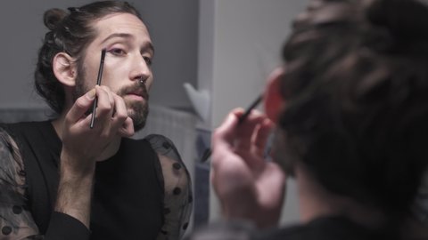 Man reflected in the mirror while applying eyelash and eyeshadow and cosmetic make-up. Real trans LGBT person primping in the bathroom at home. Diversity Transgender and Gay Concept