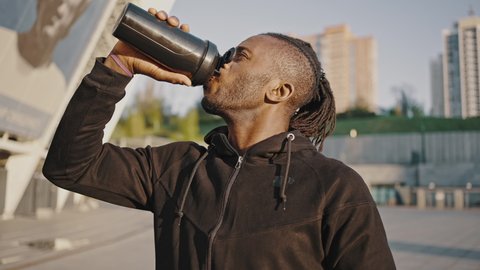 Weight gain diet. African american man athlete mixing protein cocktail in sport shaker and drinking it, practicing sports workout outdoors, close up, tracking shot
