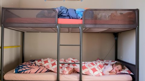 Preschoolers brother and sister lie on grey metal bunk bed under different coloured blankets in narrow children room closeup