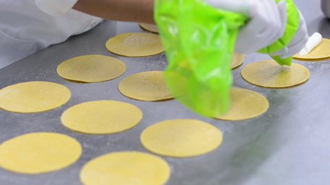 Woman with nylon gloves on, using a pastry bag to squeeze a cheese stuffing onto pasta circles, before folding them up.