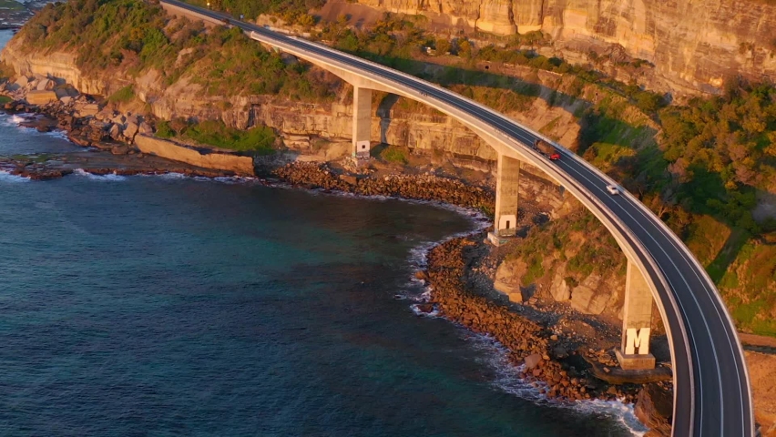 Vehicles Travelling At Sea Cliff Bridge Over Calm Blue Ocean At Sunrise In New South Wales, Australia. - aerial Royalty-Free Stock Footage #1073428199