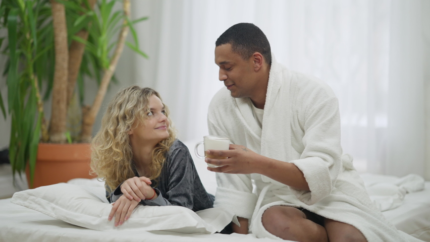 African American loving man bringing morning coffee for Caucasian woman in bed. Wide shot portrait of happy smiling boyfriend taking care of girlfriend in bedroom at home. Love and romance