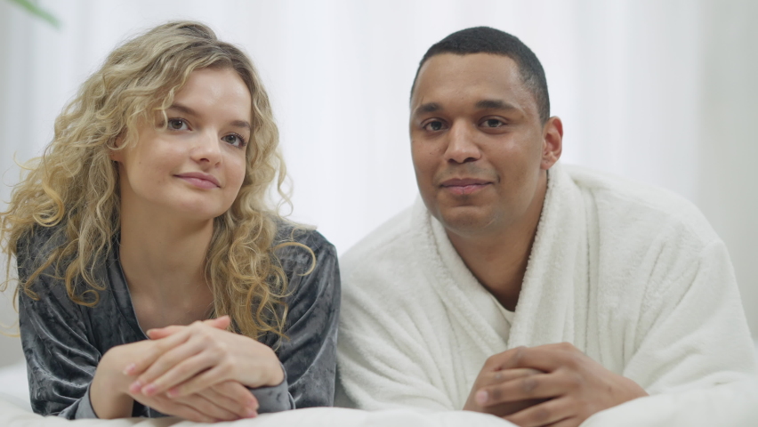 Happy loving Caucasian woman kissing African American smiling man on cheek laughing. Portrait of happy beautiful slim girlfriend in love expressing feelings to cheerful boyfriend lying on bed