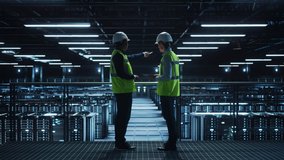 Data Center IT Specialist and System administrator Talk, Use Tablet Computer, Wearing Safety West. Server Cloud Farm Facility with Two Information Technology Engineers checking Cyber Security. Wide - Powered by Shutterstock - Get 15% off with code: PIKWIZARD15