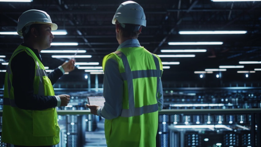 Data Center IT Specialist and System administrator Talk, Use Tablet Computer, Wearing Safety Wests. Server Clod Farm with Two Information Technology Engineers checking Cyber Security. Medium Wide Royalty-Free Stock Footage #1073440160