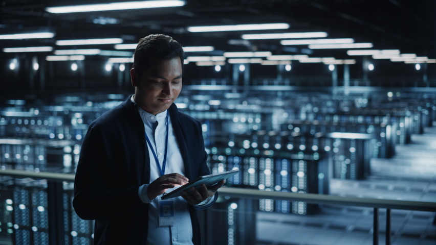Handsome Smiling IT Specialist Using Tablet Computer in Data Center, Looking at Camera. Succesful Businessman and e-Business Entrepreneur Overlooking Server Farm Cloud Computing Facility. Medium Wide Royalty-Free Stock Footage #1073440184