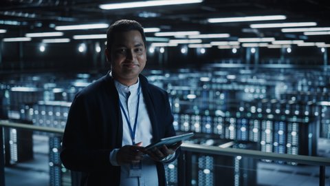 Handsome Smiling IT Specialist Using Tablet Computer in Data Center, Looking at Camera. Succesful Businessman and e-Business Entrepreneur Overlooking Server Farm Cloud Computing Facility. Medium Wide