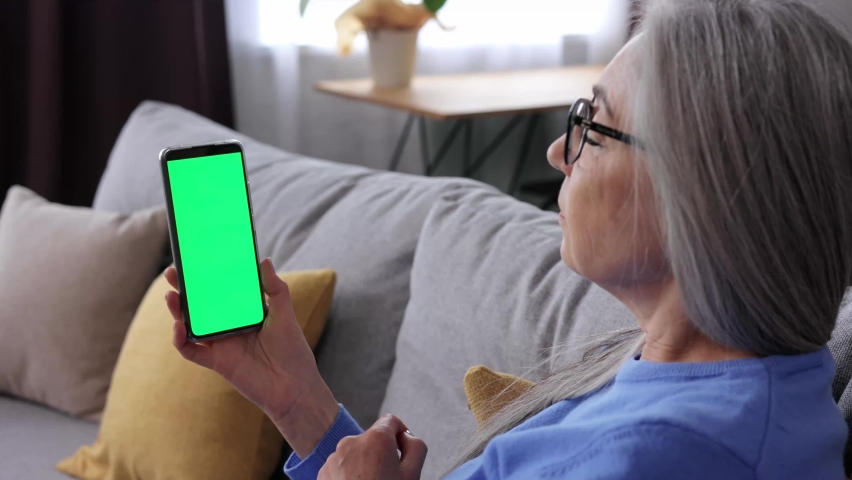 Elderly gray-haired woman making video call at smartphone camera green screen. Senior lady talking and listening during video chat with friends or colleagues. Over shoulder mobile phone screen view.