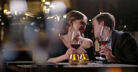 Caucasian guy leaning closer to talk to his girlfriend during a date. Couple in love drinking red wine during a romantic dinner close up 4k footage