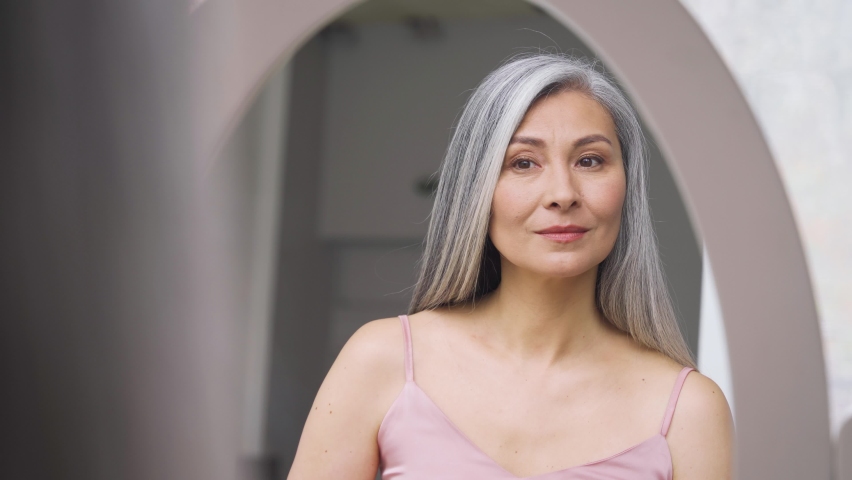 Senior older middle aged Asian woman with grey hair holding make up brush on radiant face with perfect skin looking at mirror smiling to her reflexion. Ads of makeup foundation for natural glow skin. Royalty-Free Stock Footage #1073456330