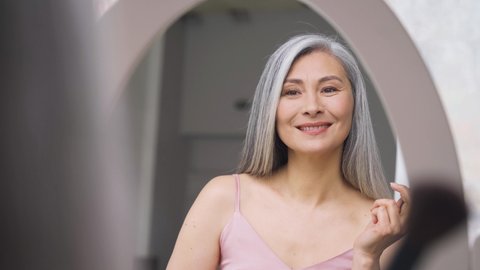 Senior older middle aged Asian woman with grey hair holding make up brush on radiant face with perfect skin looking at mirror smiling to her reflexion. Ads of makeup foundation for natural glow skin.