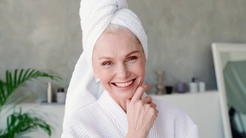 Portrait of happy smiling attractive middle aged woman wearing bathrobe and white towel touching skin winking looking at camera. Advertising of bodycare spa procedures concept.