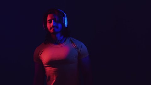 Music lover. Attractive dancing man. Neon light portrait. Stereo technology. Smiling guy in wireless headphones moving in rhythm of sound on dark blue red color glow copy space.