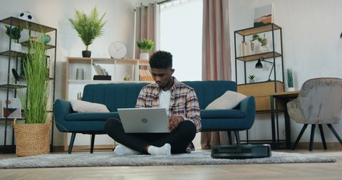 Smart home concept where good-looking emotional 20-aged african american guy sitting on the floor near couch at home and working on laptop while automatic vacuum cleaner cleaning carpet