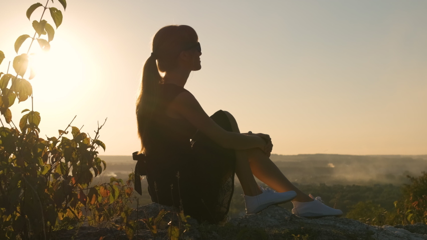 Young pretty woman in black short summer dress sitting on a rock relaxing outdoors at sunset. Fashionable female enjoying warm evening in nature.