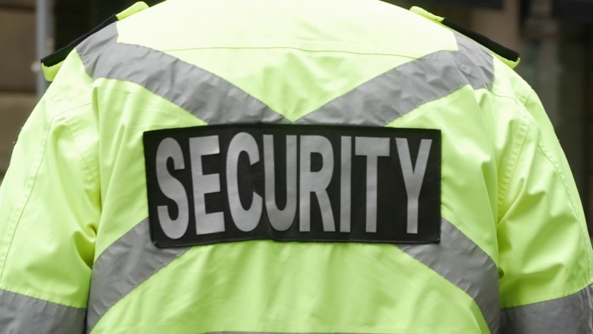 A rearview of security personnel in a neon-green safety jacket walking along the street in HD