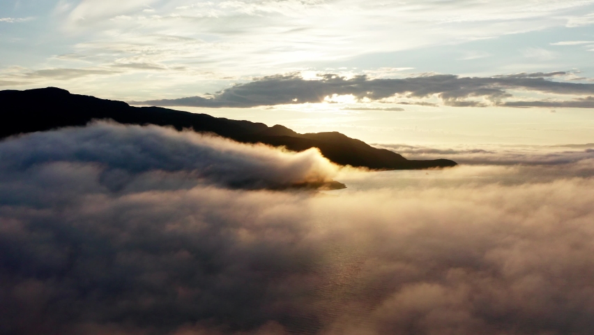 Drone Flying Above The Clouds Over The Sea With Rocky Mountain In Silhouette On A Golden Hour Sunrise - wide aerial shot Royalty-Free Stock Footage #1073475236