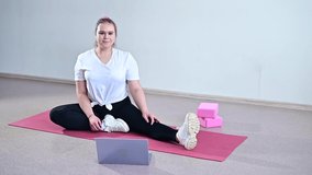 Young caucasian fat woman doing bends on a sports mat and watching a training video on a laptop. A chubby girl doing stretching remotely using video communication