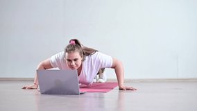 A young obese woman does push-ups. The girl is engaged in fitness at home on the mat and watches a training video on a laptop
