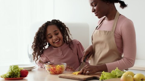 Young african american mother preparing vegetable salad, curly little daughter coming and eating fresh pepper at kitchen, running away to play, slow motion